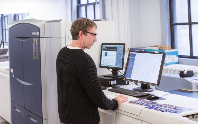 A Copytech staff member stands while working in front of two large monitors and a printer.