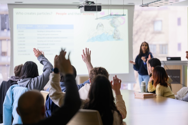 Classroom scene with students raising their hands toward a projector screen, while an instructor stands at the front engaging with them.