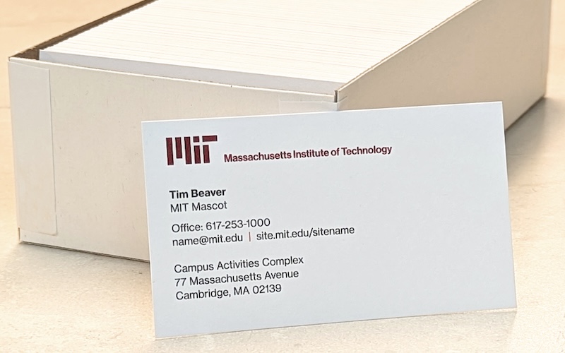 A business card using the MIT standard template rests next to a box full of business cards.