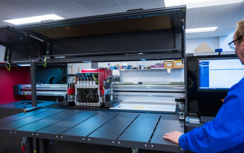 A Copytech staff member operates a large printer with its top cover open, revealing internal components. They are standing beside the printer, looking at a computer screen displaying printing-related data.